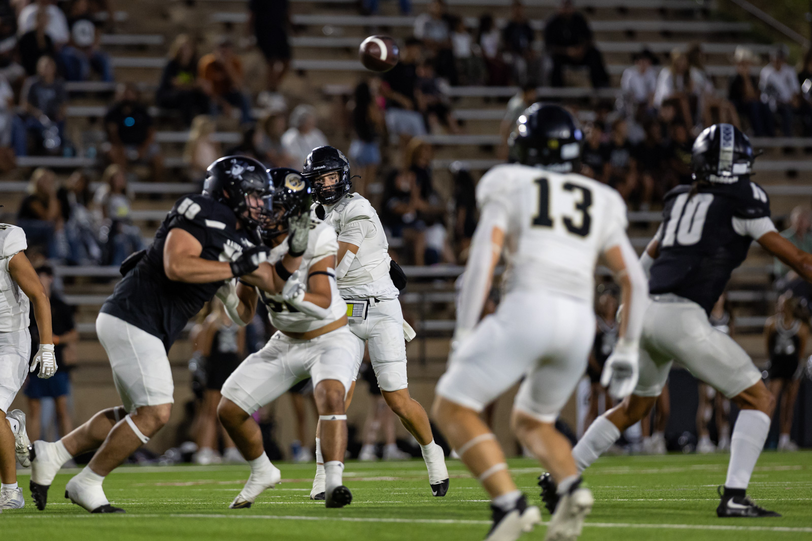 CHS-RHS-FB-16 | Press Pass Sports White-uniform quarterback surveys the field as defenders collide and the ball sails through the air behind them; night game.