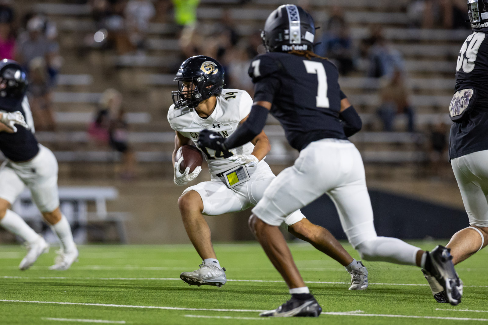 CHS-RHS-FB-17 | Press Pass Sports A football player in white jersey runs with the ball as black-clad defenders chase him on a green turf field at a night game.