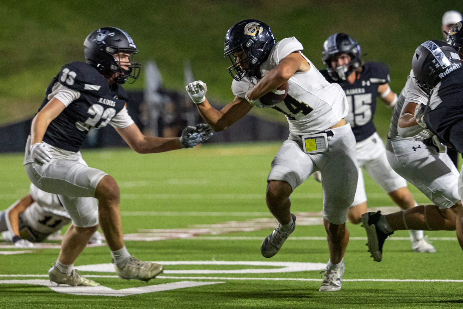 CHS-RHS-FB-18 | Press Pass Sports Running back in white carries the ball as a defender in black closes in on a night football field, dodging the tackle.