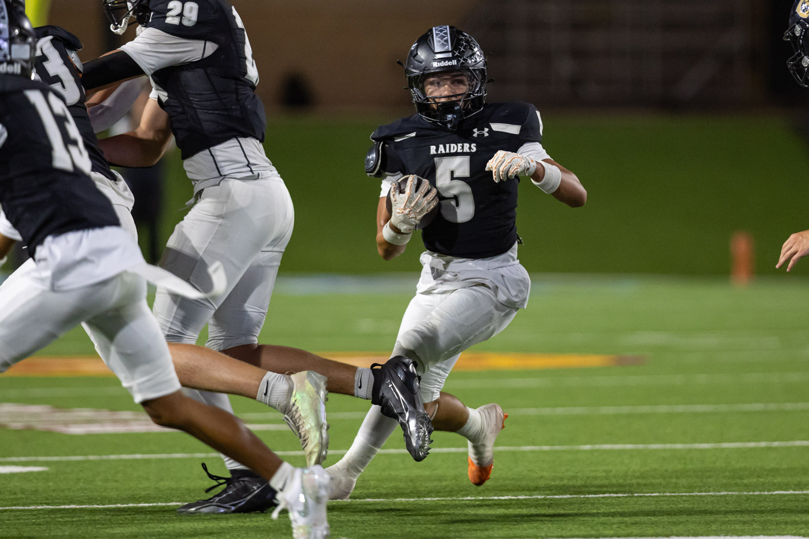 CHS-RHS-FB-23 | Press Pass Sports Football player with number 5 Raiders runs with the ball while tackling opponents nearby on a green field at night