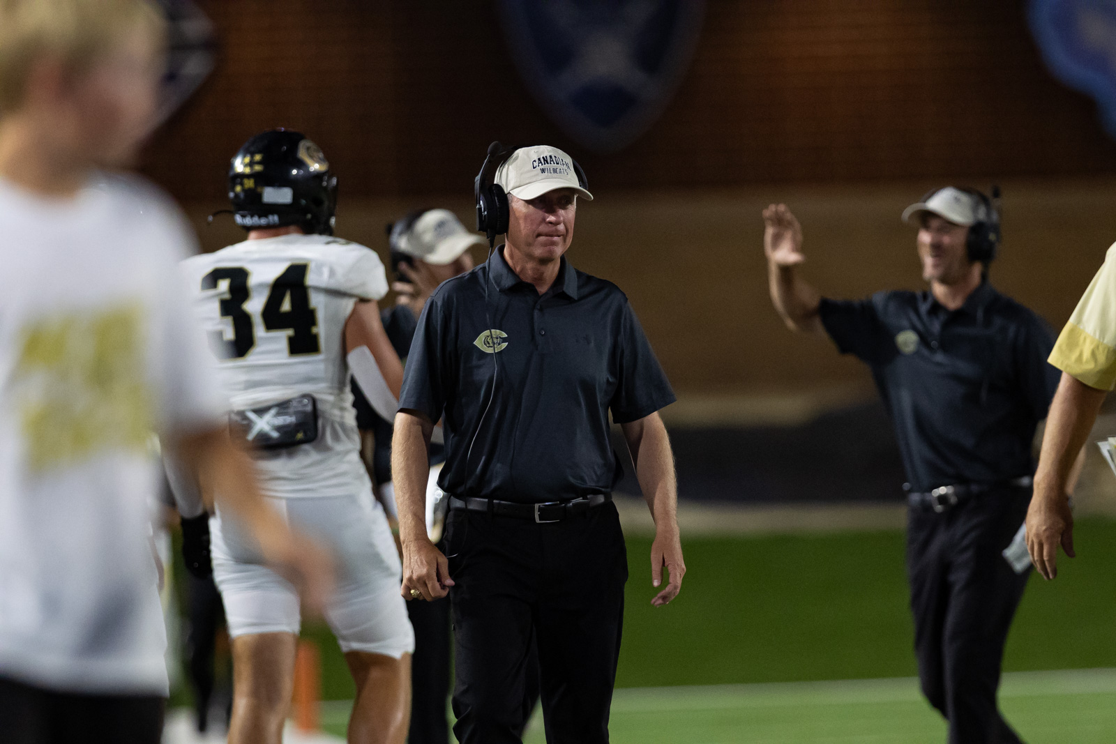 CHS-RHS-FB-25 | Press Pass Sports Football coach wearing a headset and cap on the sideline as players stand nearby.