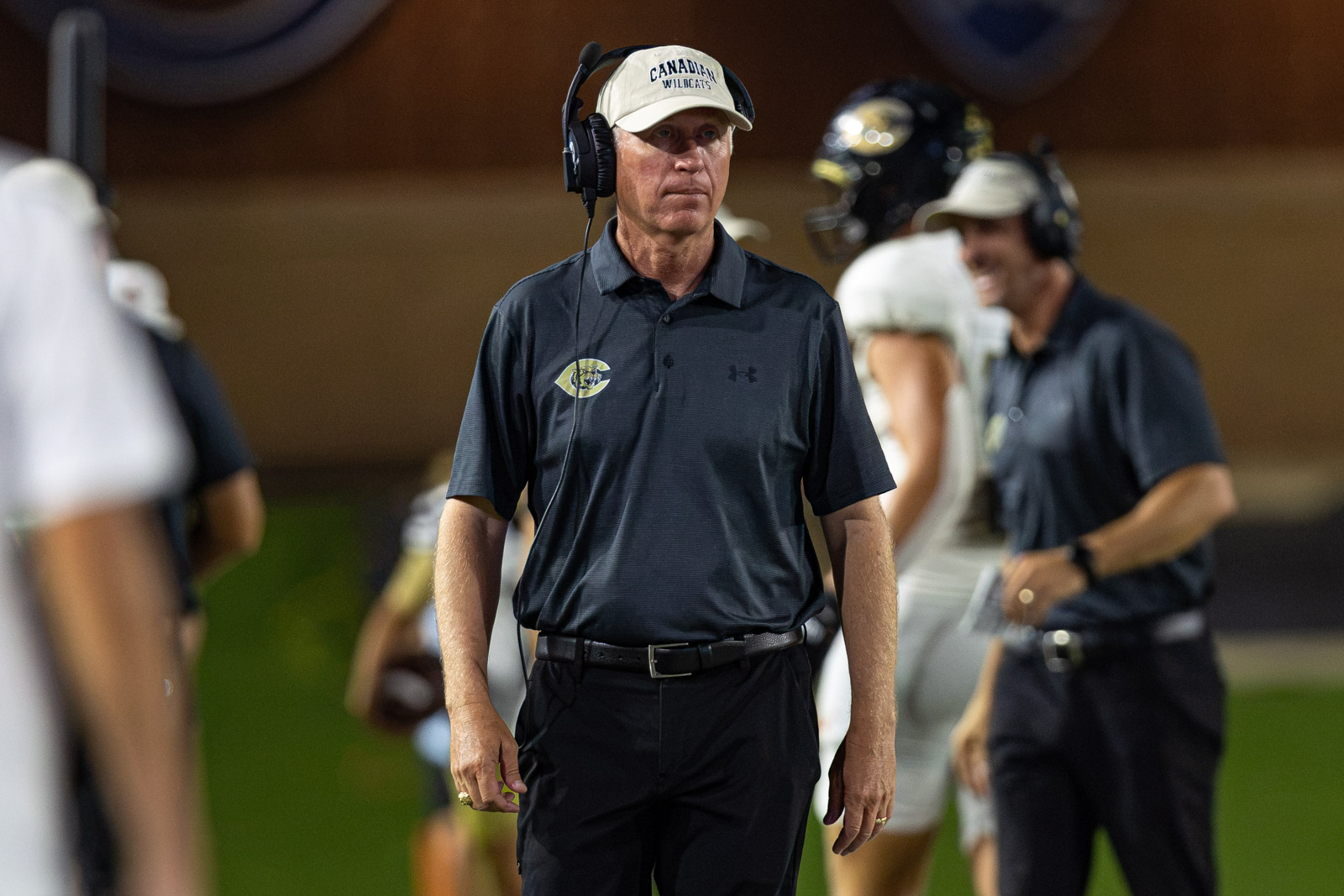 CHS-RHS-FB-26 | Press Pass Sports Male football coach wearing a headset, dark polo and cap, walking on the sideline during a game or practice.