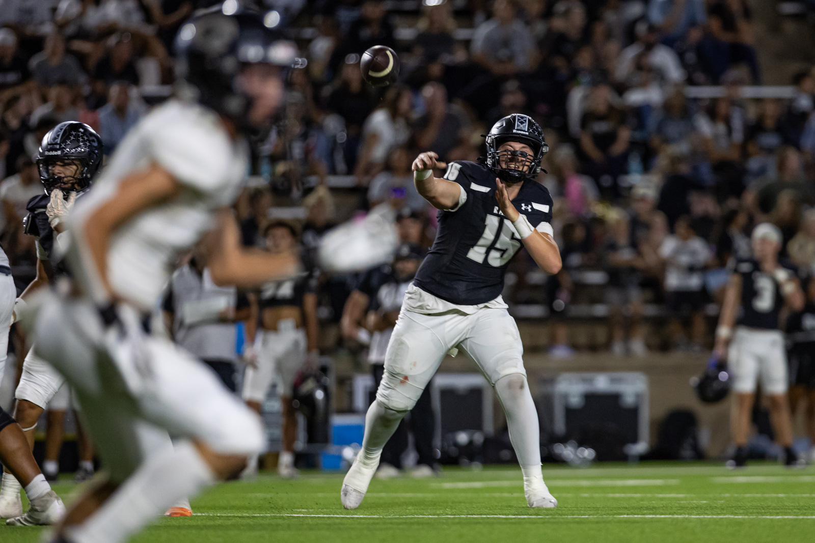 CHS-RHS-FB-27 | Press Pass Sports Quarterback in dark jersey (#15) throws a pass during a nighttime football game with a crowd in the stands behind him.