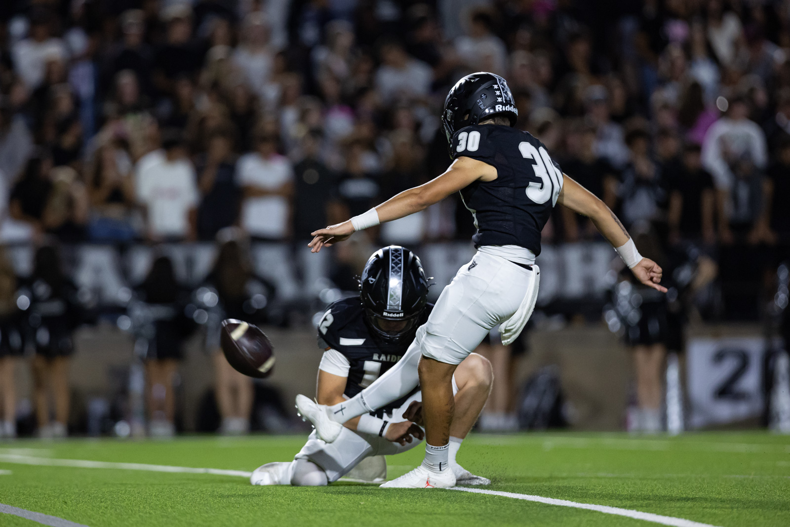 CHS-RHS-FB-30 | Press Pass Sports Football players in black uniforms on a green turf; one kicks while another dives for the ball amid a cheering crowd at night stadium.