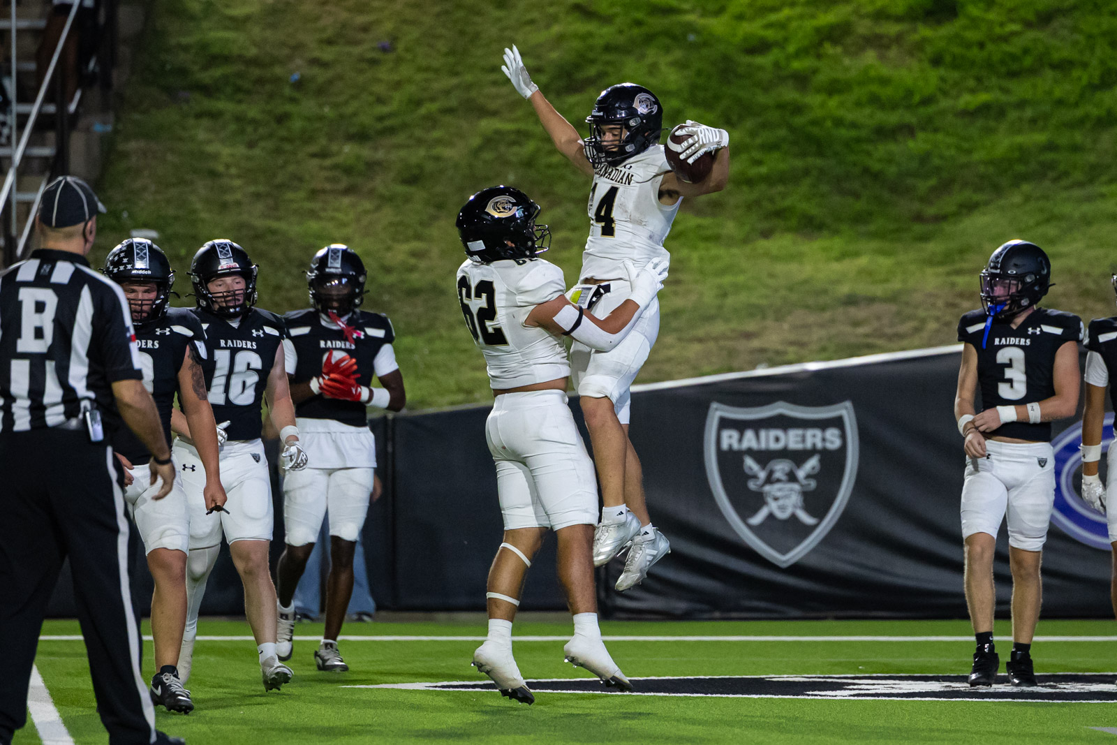 CHS-RHS-FB-35 | Press Pass Sports Football players in black Raiders uniforms celebrate as a white-uniform teammate leaps in the air with the ball during a game on a green turf field with a Raiders banner in the background.