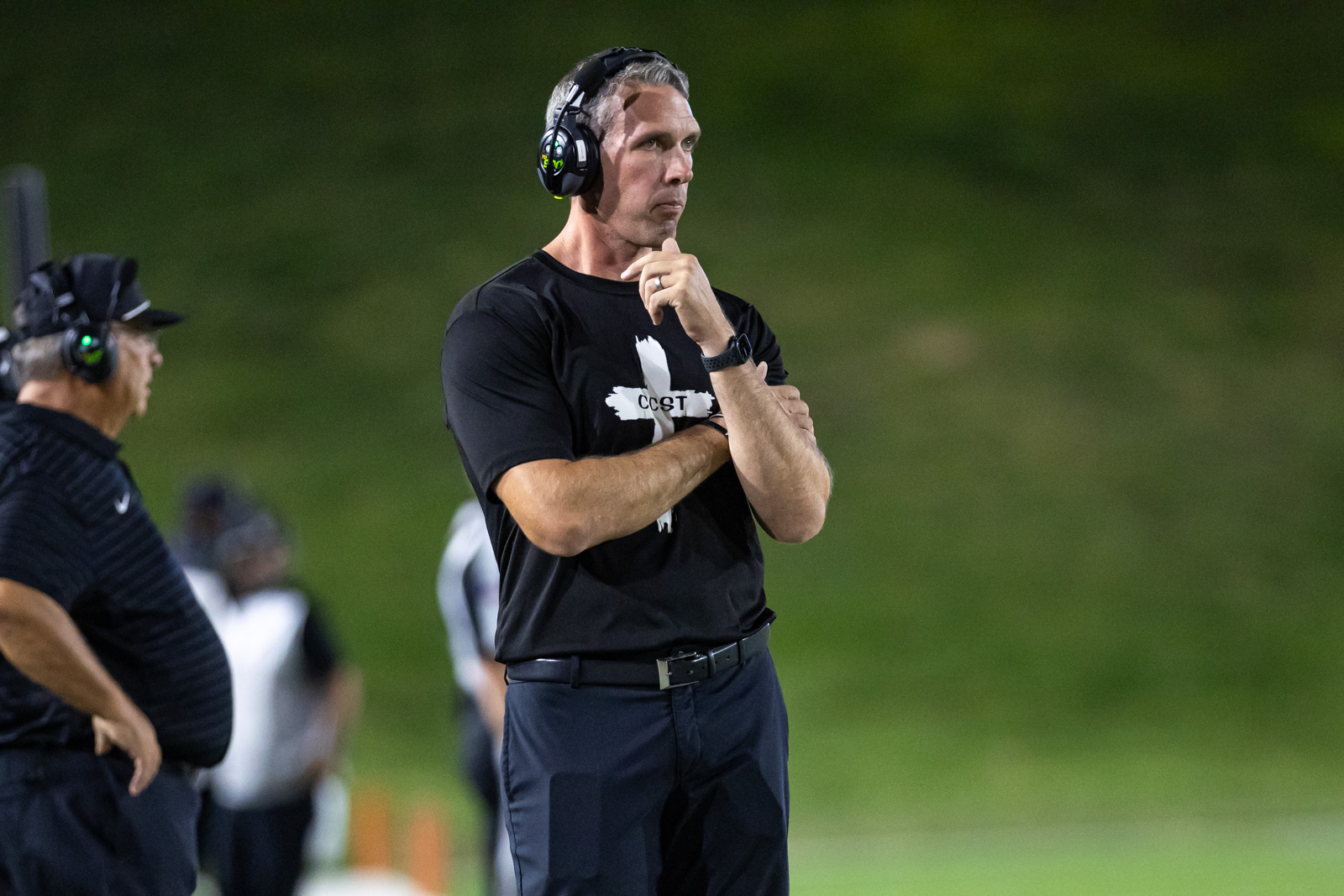 CHS-RHS-FB-37 | Press Pass Sports Coach with headset in black shirt stands on sideline, hand on chin, deep in thought during a game.