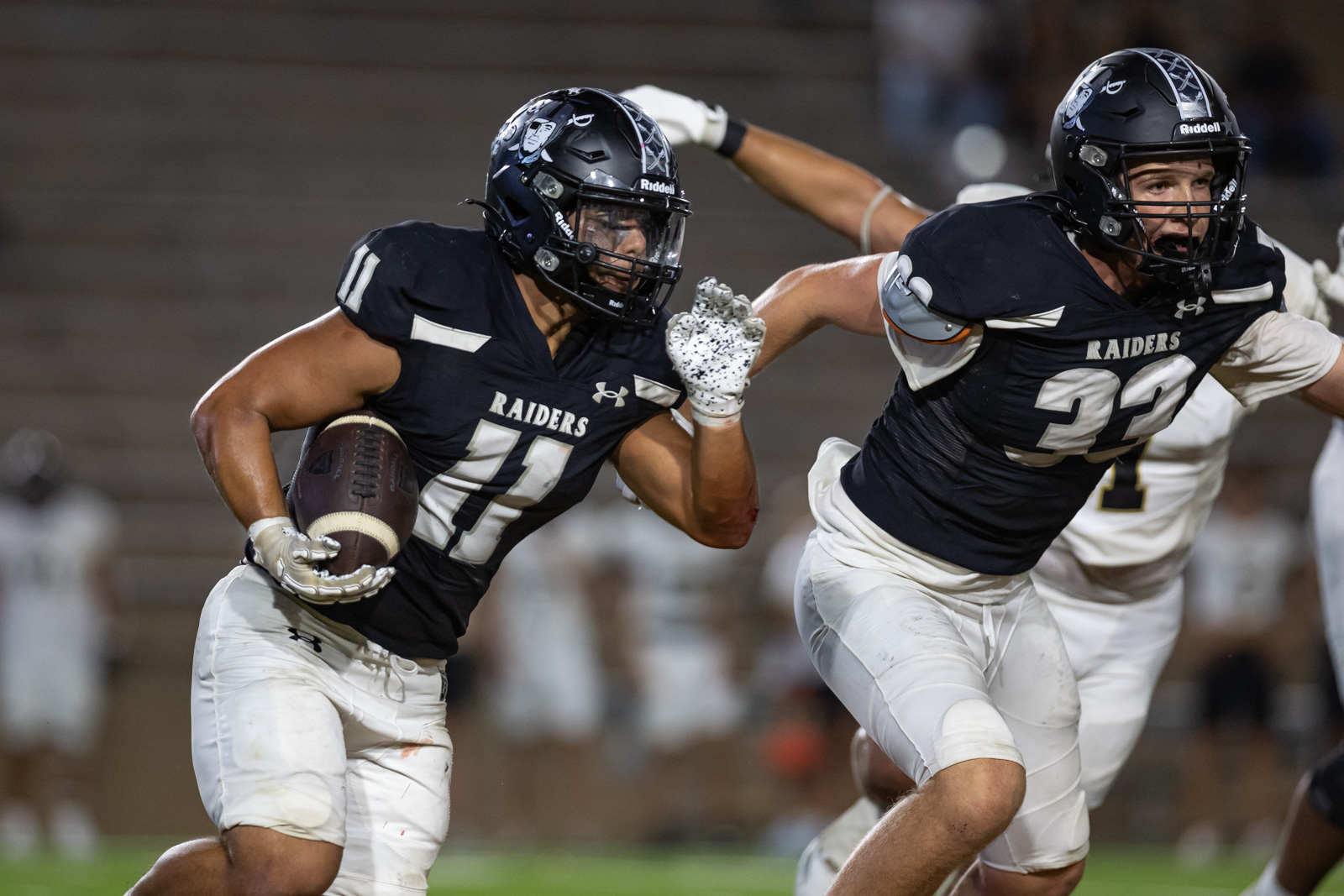 CHS-RHS-FB-41 | Press Pass Sports Raiders running back #11 carries the ball as he runs forward, with teammate #21 close behind in pursuit on a night game.
