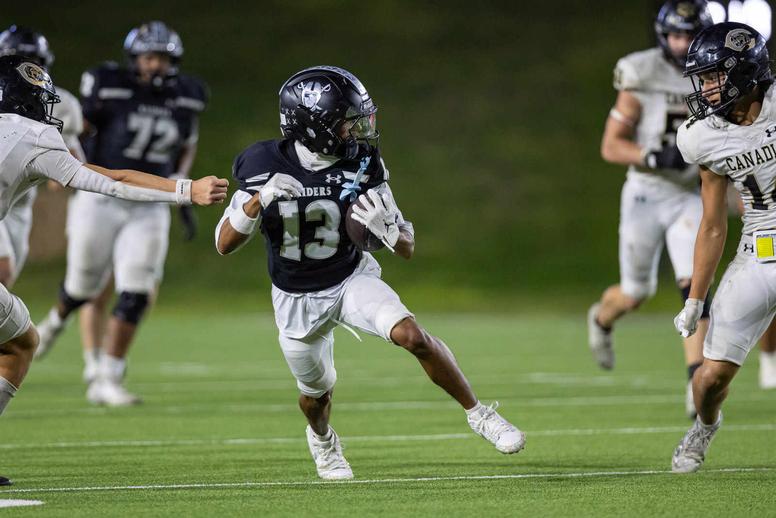 CHS-RHS-FB-44 | Press Pass Sports Running back wearing number 13 in a dark Raiders jersey runs with the football as defenders close in on a green turf field at night