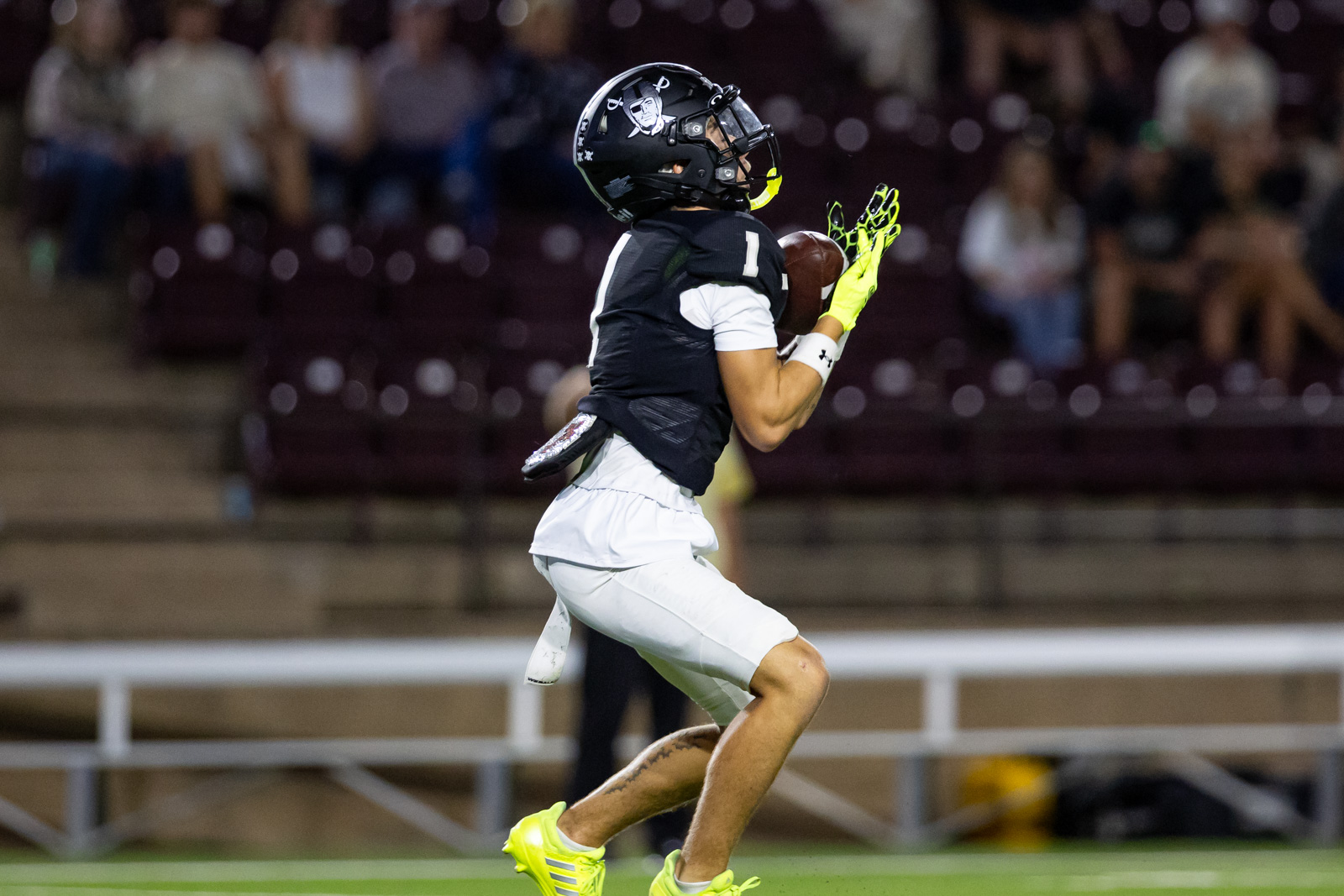 CHS-RHS-FB-46 | Press Pass Sports Football player in black jersey and white shorts catches a football in midair, neon gloves on, stadium stands in background.