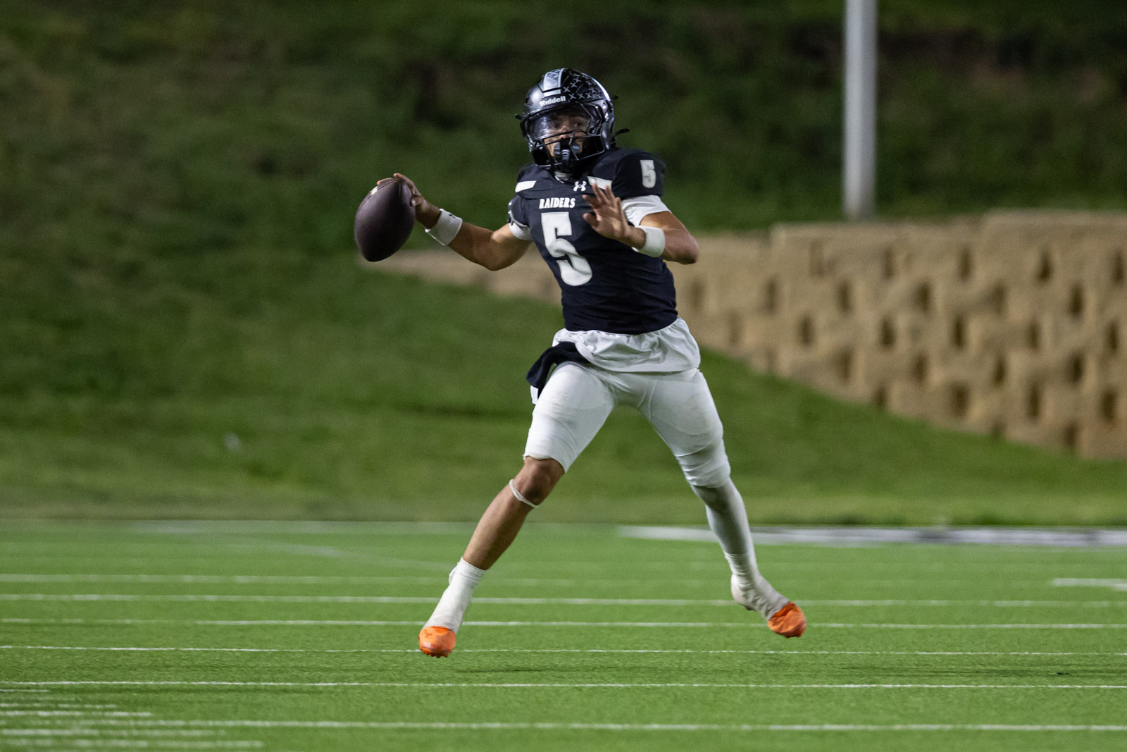 CHS-RHS-FB-47 | Press Pass Sports Raiders quarterback wearing number 5 runs with a football on a green turf field at dusk/dark, looking to pass or evade defenders