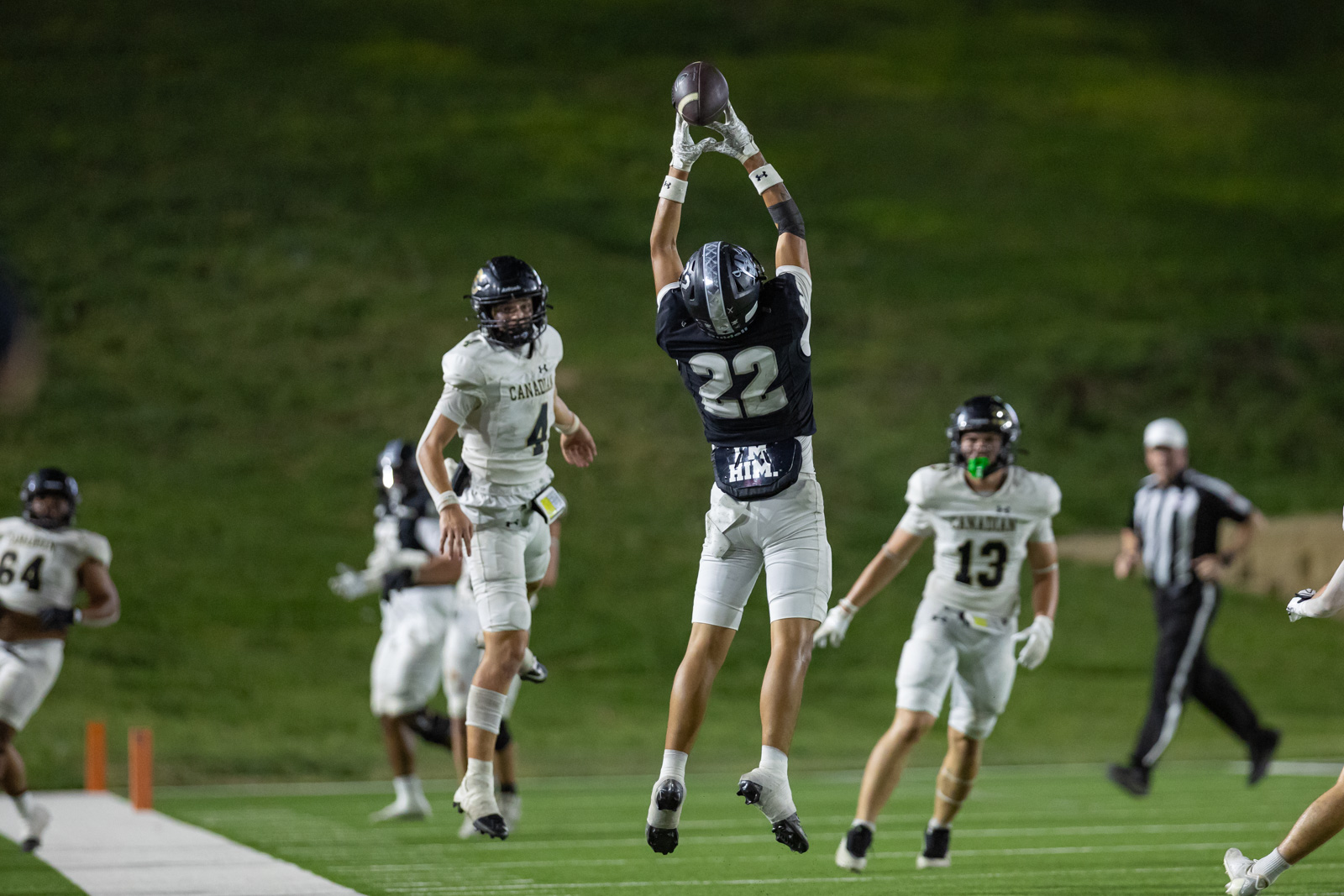 CHS-RHS-FB-48 | Press Pass Sports A football player in dark jersey (number 22) leaps with arms raised to catch a ball, others in white jerseys nearby on a green field at night coat.