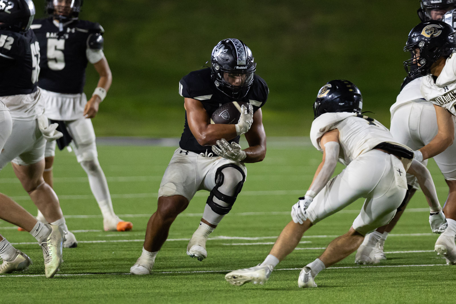 CHS-RHS-FB-49 | Press Pass Sports Football player in a dark uniform runs with the ball, dodging white-uniform defenders on a bright turf field under night lights.