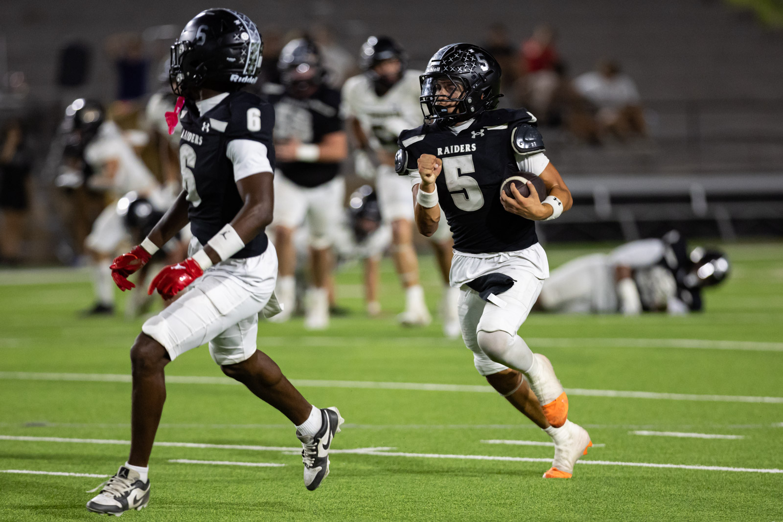 CHS-RHS-FB-50 | Press Pass Sports Number 5 Raiders running with the ball during a night game, black uniforms and orange cleats visible on green turf.