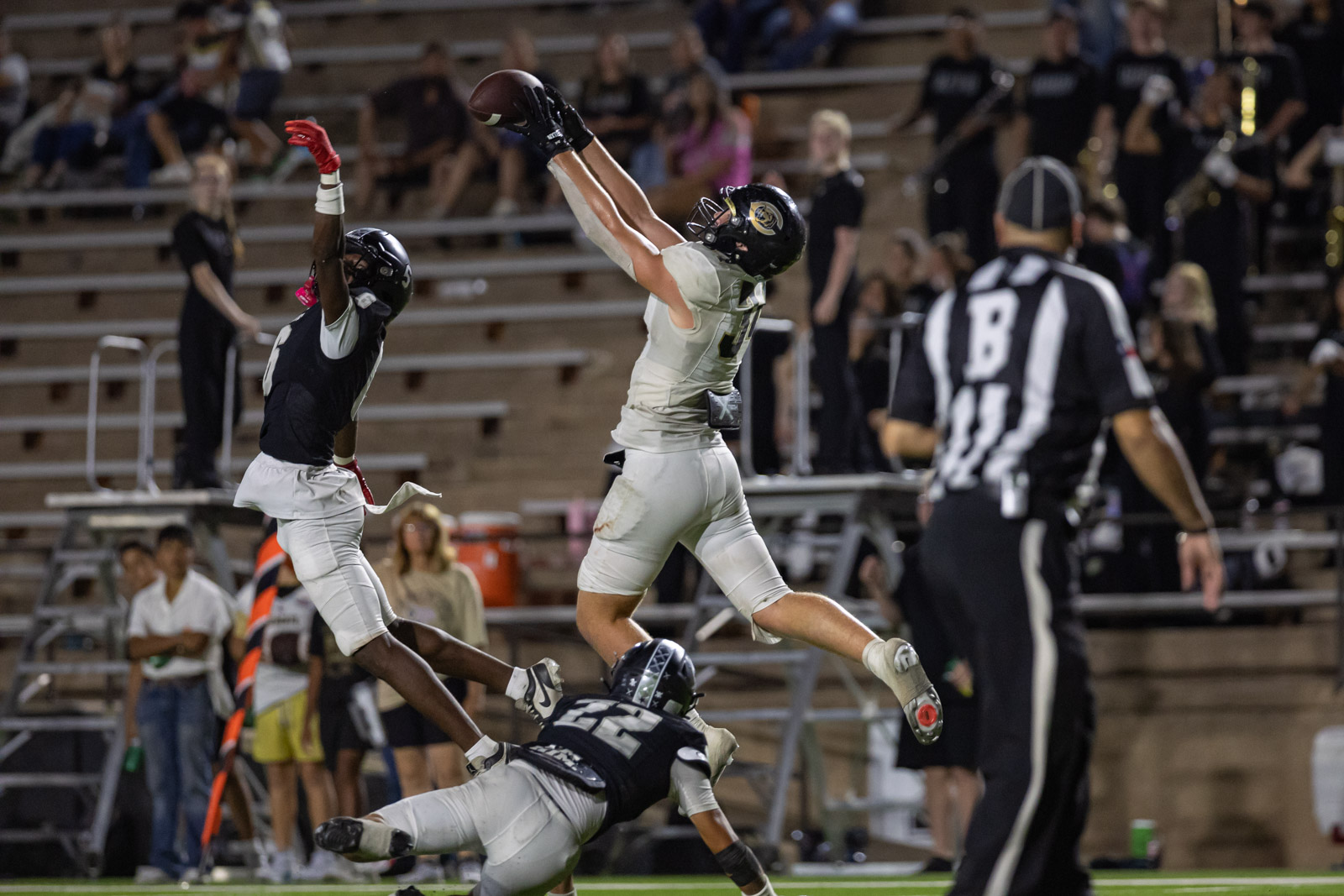 CHS-RHS-FB-54 | Press Pass Sports Players leap for a football, reaching for a catch above a defender as a third player falls near the end zone with a ref watching in the background.