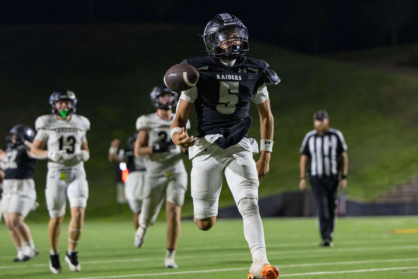 CHS-RHS-FB-57 | Press Pass Sports Football player in black Raiders jersey #5 runs with the ball during a night game, teammates in white behind him, referee in stripes in the background.