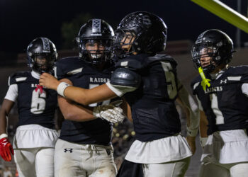 Football players in black Raiders uniforms celebrate on a night field, embracing each other after a play.