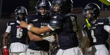 Football players in black Raiders uniforms celebrate on a night field, embracing each other after a play.