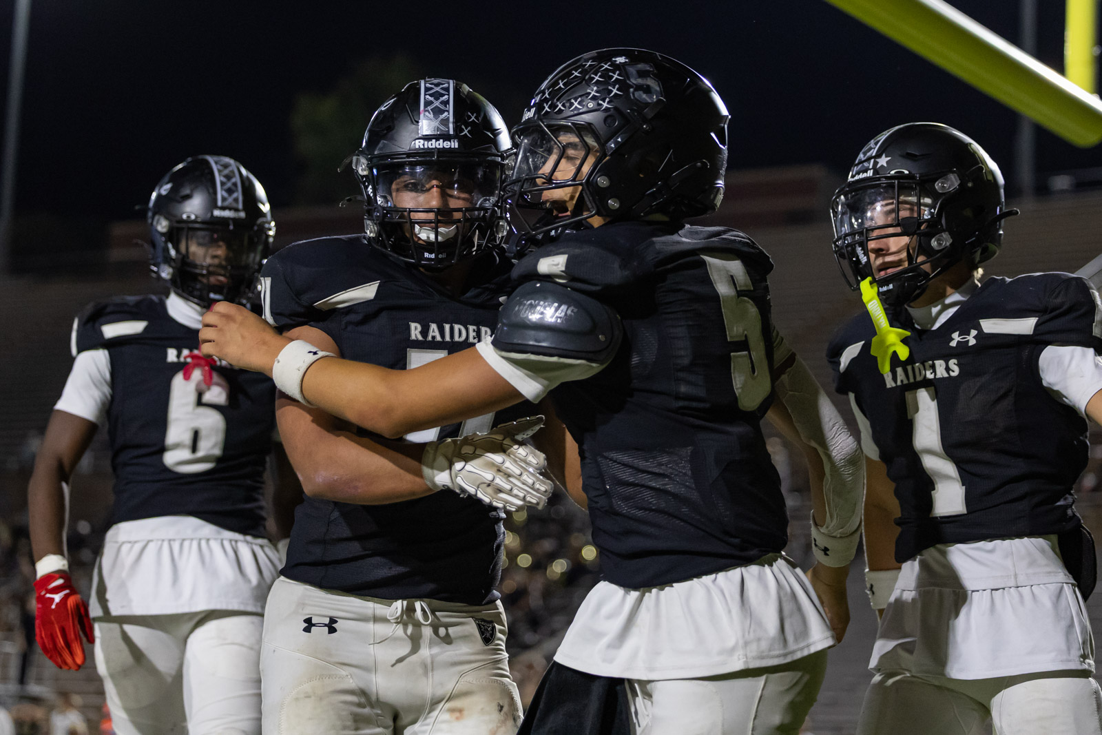 CHS-RHS-FB-58 | Press Pass Sports Football players in black Raiders uniforms celebrate on a night field, embracing each other after a play.