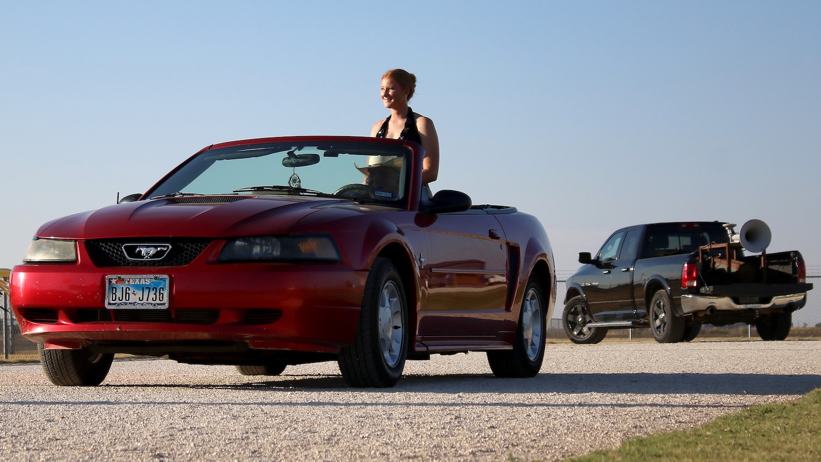 Red Ford Mustang convertible with a woman standing in the driver’s seat on a gravel road, Texas license plate visible, and a black pickup with a large loudspeaker in the background.