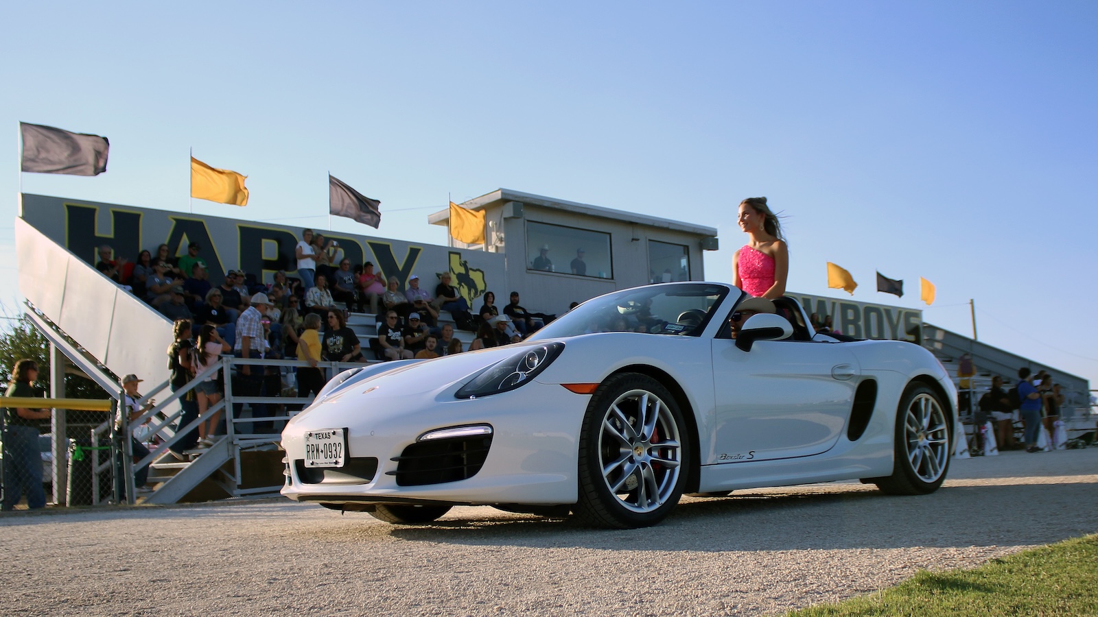 White Porsche convertible (Boxster S) with a woman in a pink dress sitting on the car, crowds in bleachers at a race or car show on a sunny day.