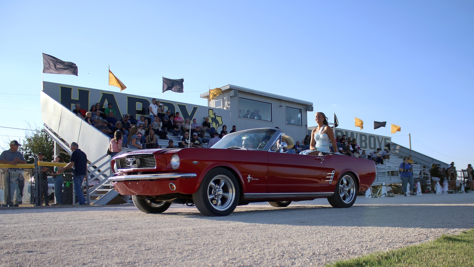 Red vintage Ford Mustang convertible drives by in a parade, a woman in a light dress sits in the passenger seat as spectators watch from bleachers behind.