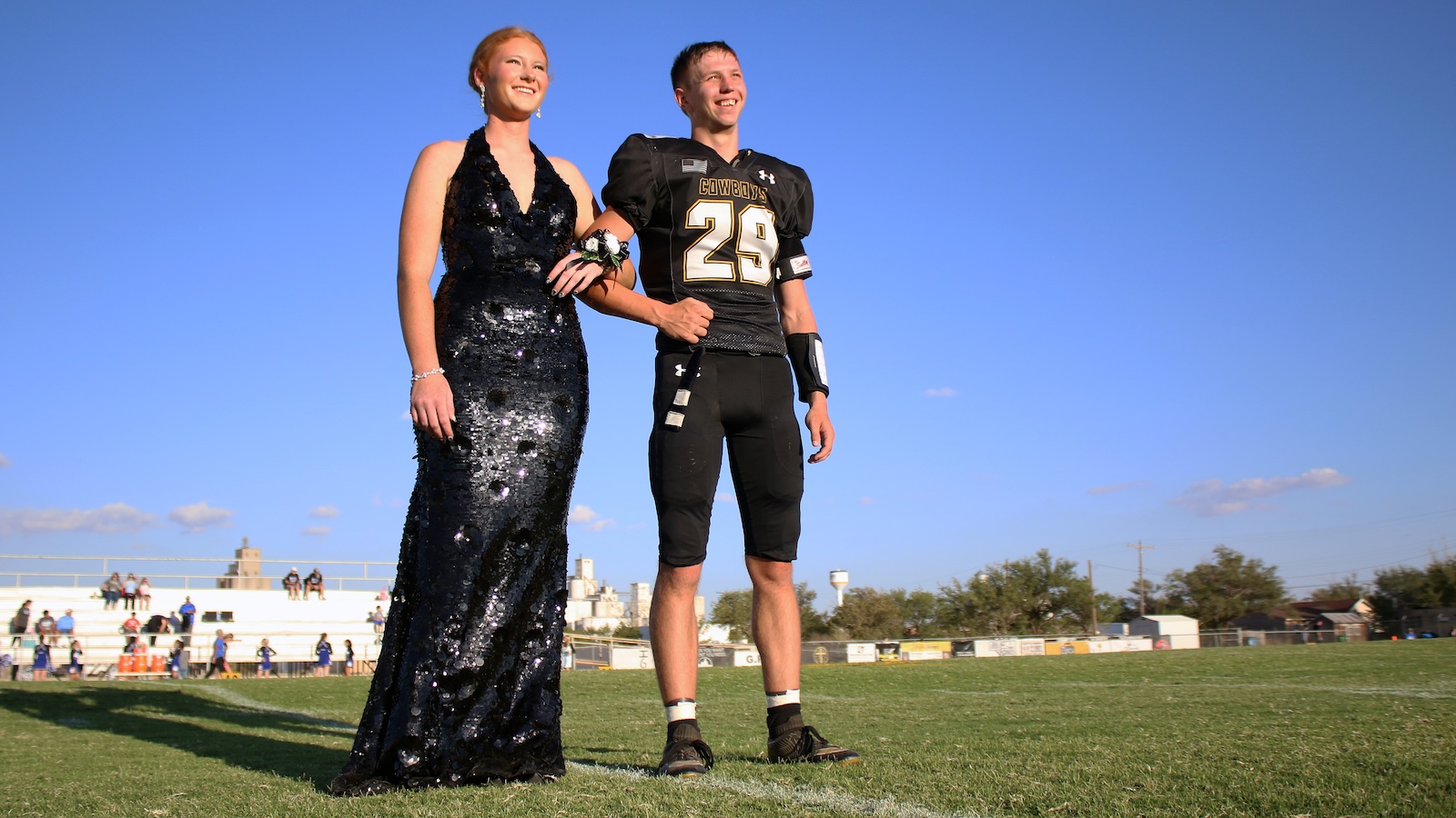 Woman in a shimmering black gown stands arm-in-arm with a football player wearing a black jersey #29 on a grassy field under a clear blue sky, both smiling.