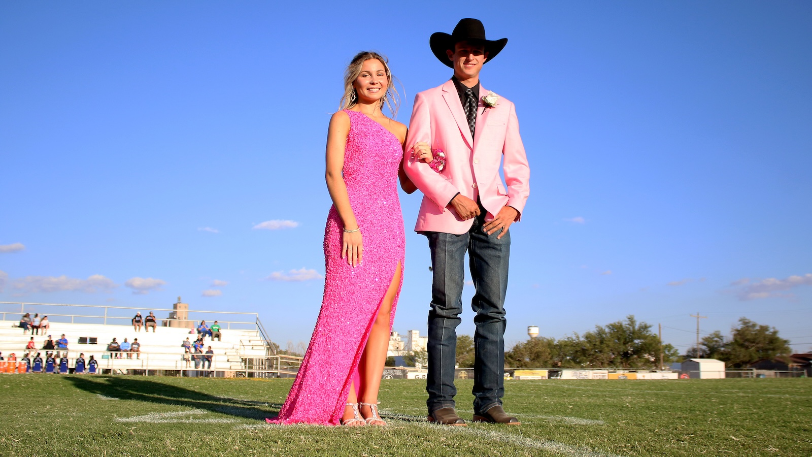 Couple posing on a sunny sports field; woman in a pink sequin gown and man in a pink blazer with a black cowboy hat, arm in arm.
