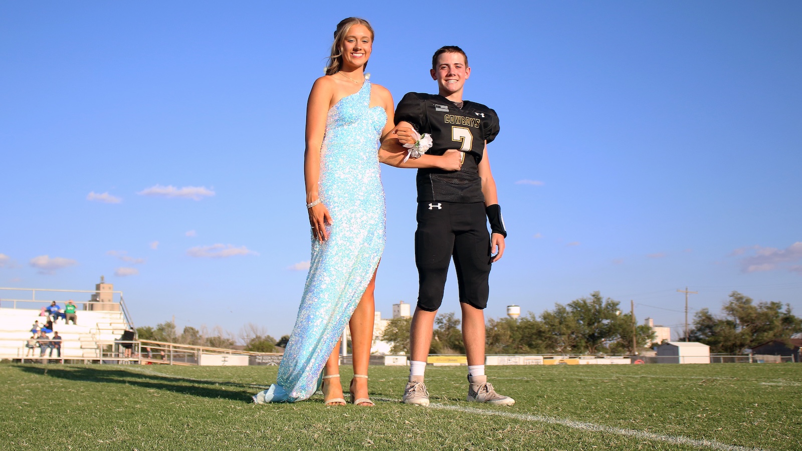 Two teenagers pose on a football field; girl in a light blue sequined dress links arms with a boy in a Cowboys uniform.