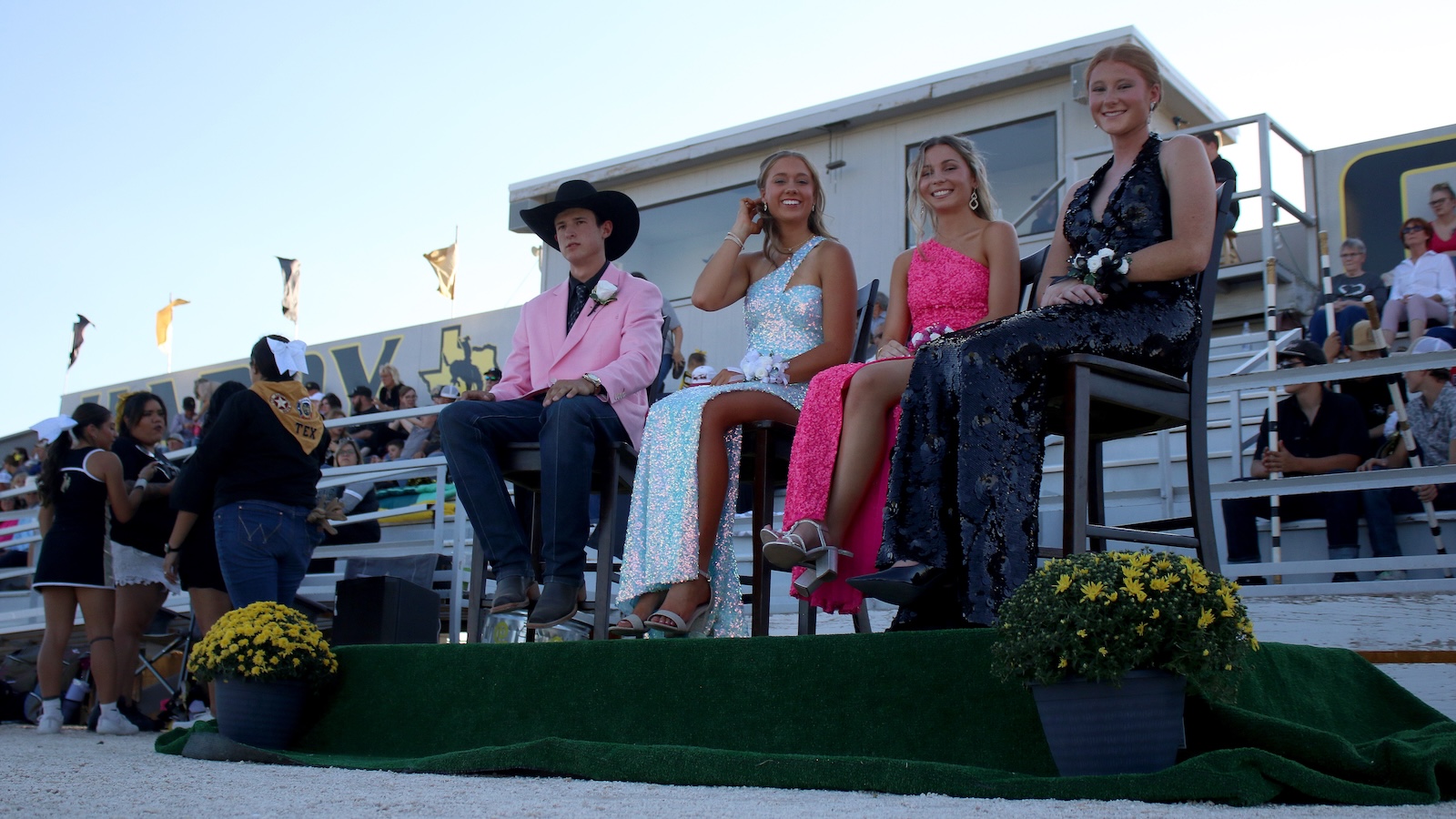 Four teens seated on a judges’ platform at a prom: a boy in a pink blazer and cowboy hat with three girls in glittery gowns nearby.