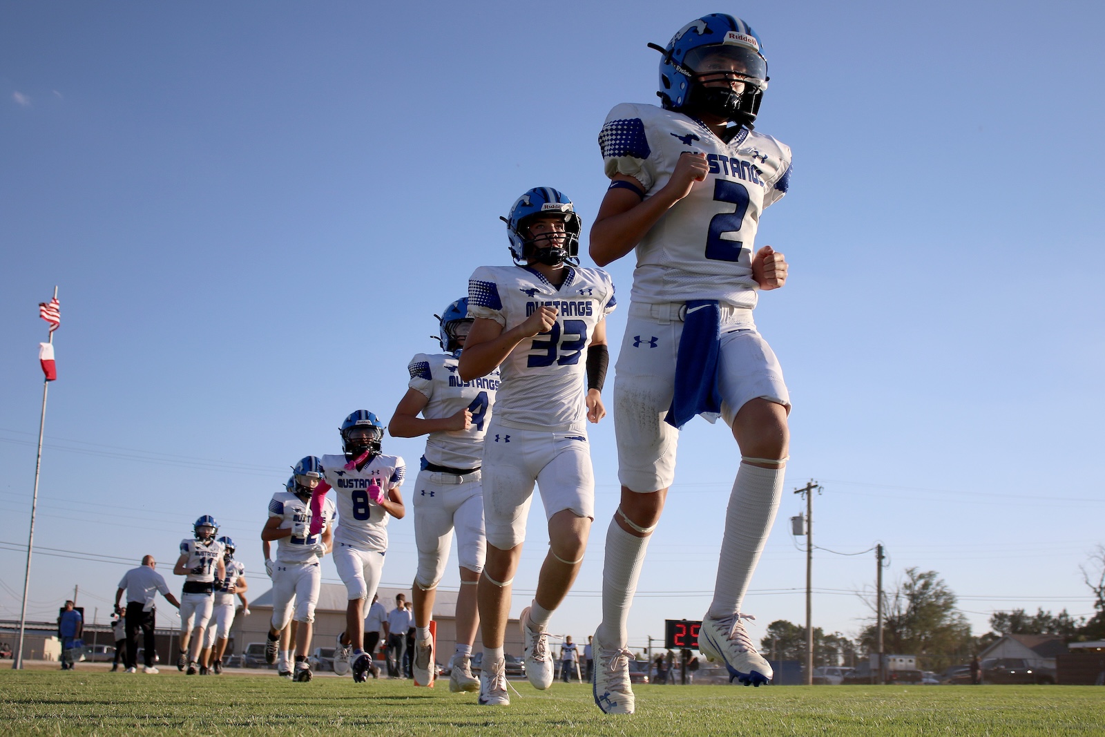 Mustangs football players sprint onto the field in white and blue uniforms under a clear blue sky.