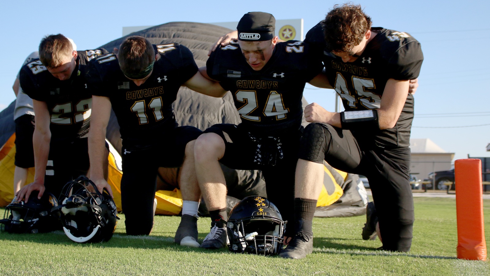 Four Cowboys players in black uniforms form a circle with arms over each other on a grassy field, a helmet resting on the ground between them.