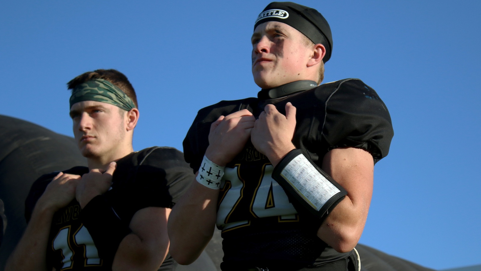 Two football players in black jerseys stand outdoors, fists clenched at their chests against a bright blue sky.