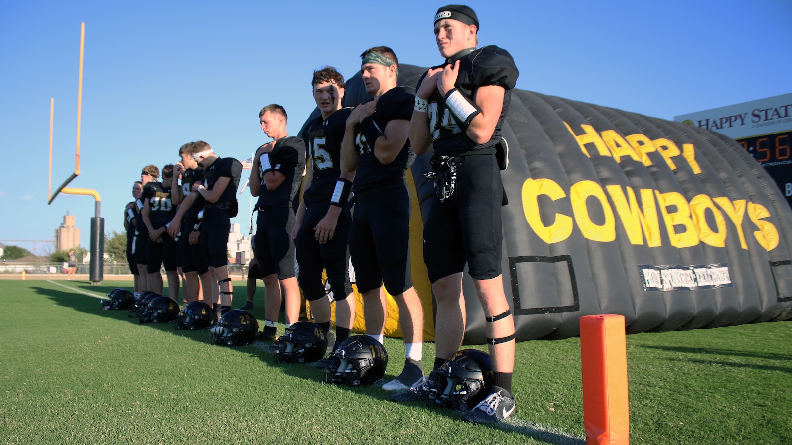 Football players lined up on a field with helmets on the grass, in front of a large black inflatable tunnel that reads'Happy Cowboys'.
