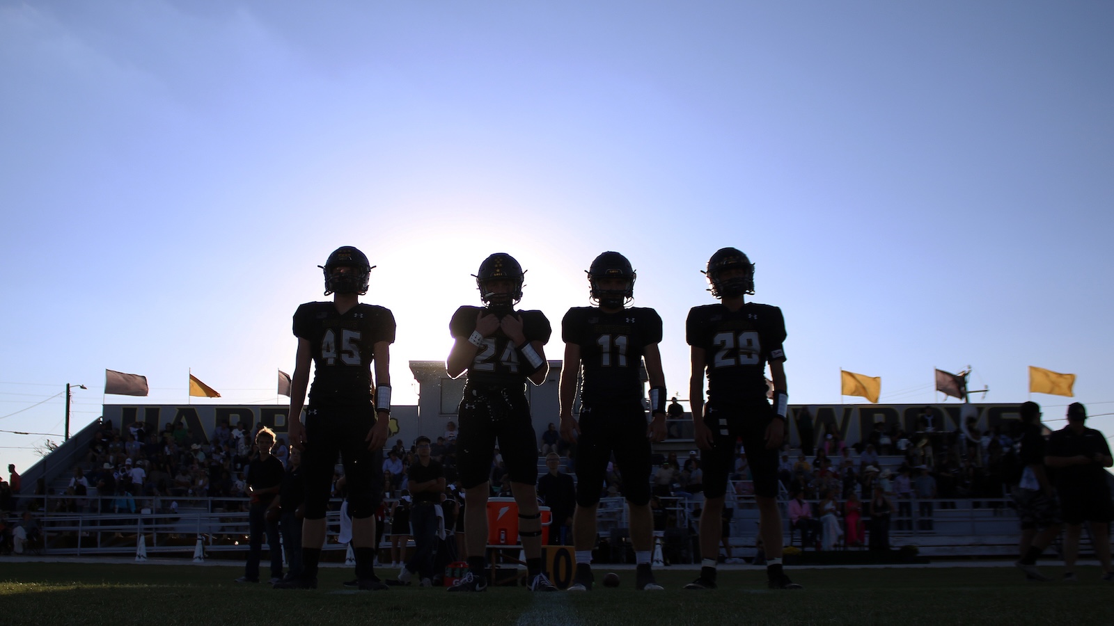 Four silhouetted football players in black uniforms stand in a row on the field with the sun behind them and a crowd in the stands behind them.