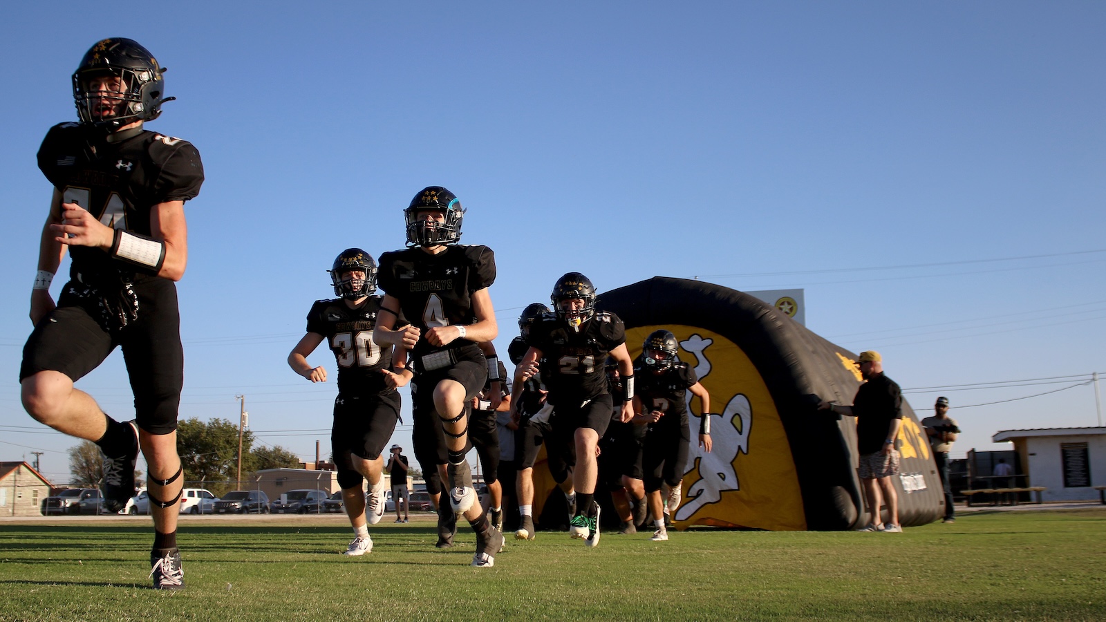 Young football players in black uniforms sprint onto the field from a yellow-and-black inflatable tunnel as a game begins.