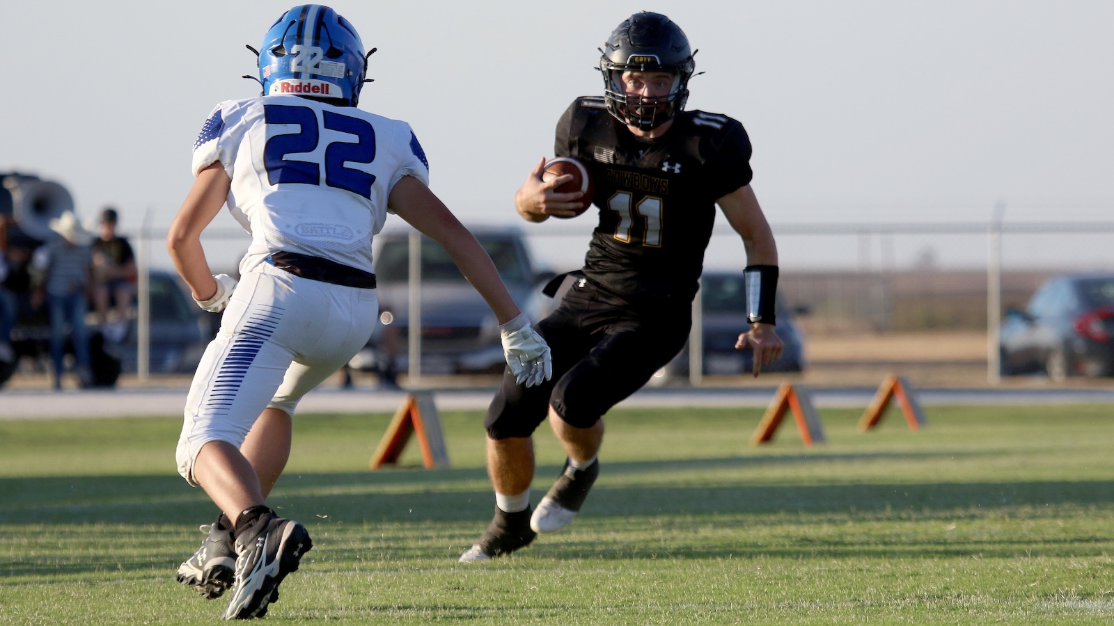 Two youth football players in action on a grassy field: a white-jersey defender (#22) approaching a black-jersey ball carrier (#11) with the ball tucked under his arm, blurred spectators and cones in the background.