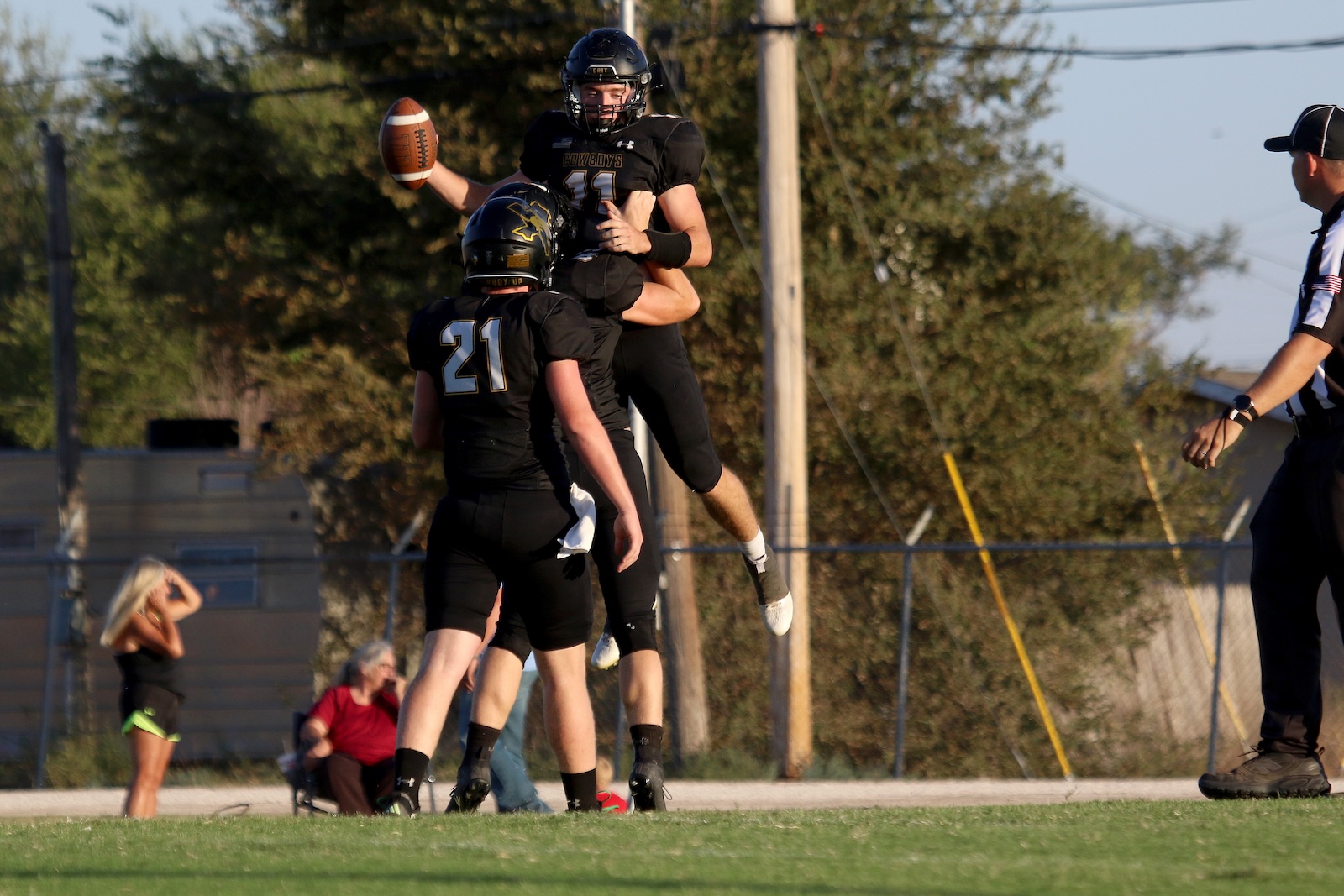 Football players in black uniforms jump for a catch as a teammate reaches up to grab the ball mid‑air on a sunny field.