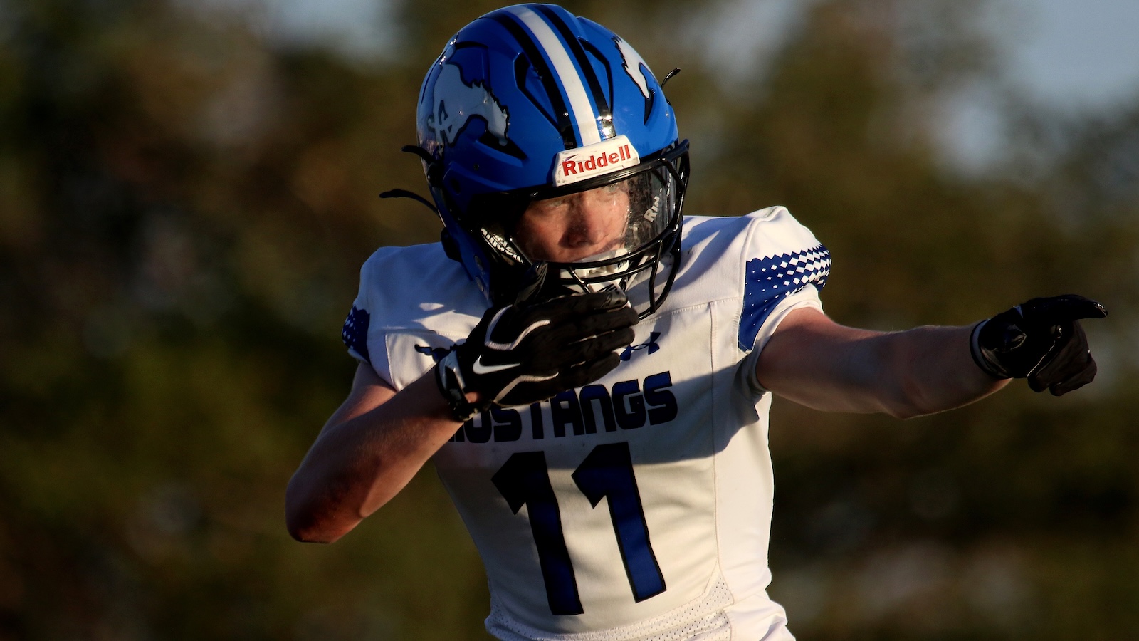Football player in a blue helmet and white jersey with number 11 points forward during play in a sunny field, Mustangs team name visible.