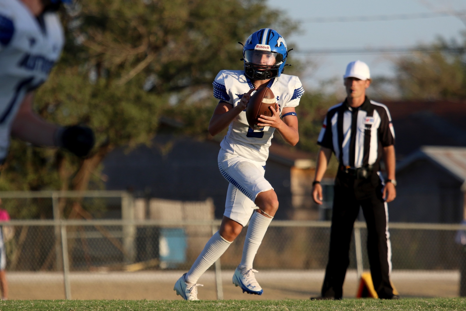 Youth football player in a blue helmet and white uniform catches a football while running on the field, with a striped referee watching in the background.