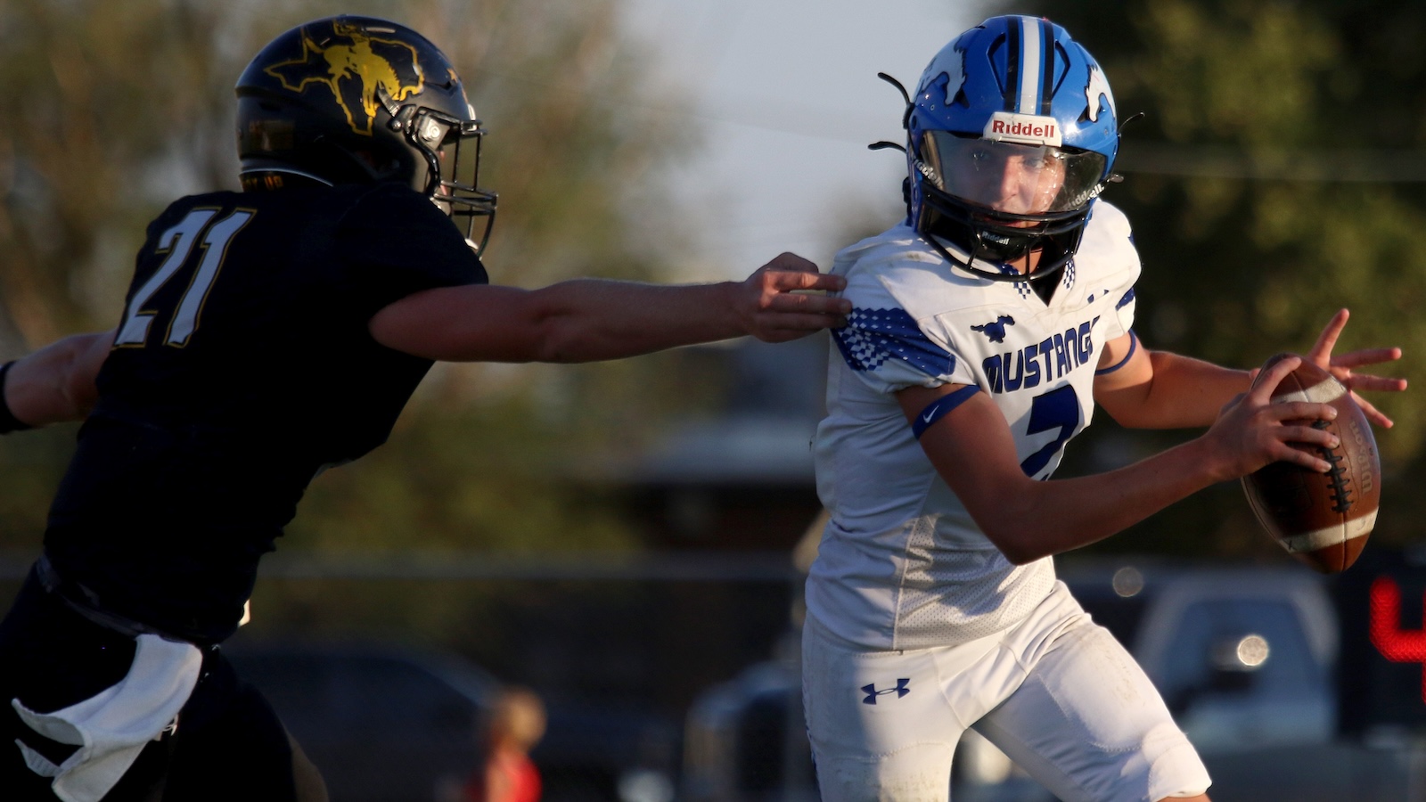 Mustangs player in white uniform (number 7) grips a football while a defender in black reaches in to tackle him.