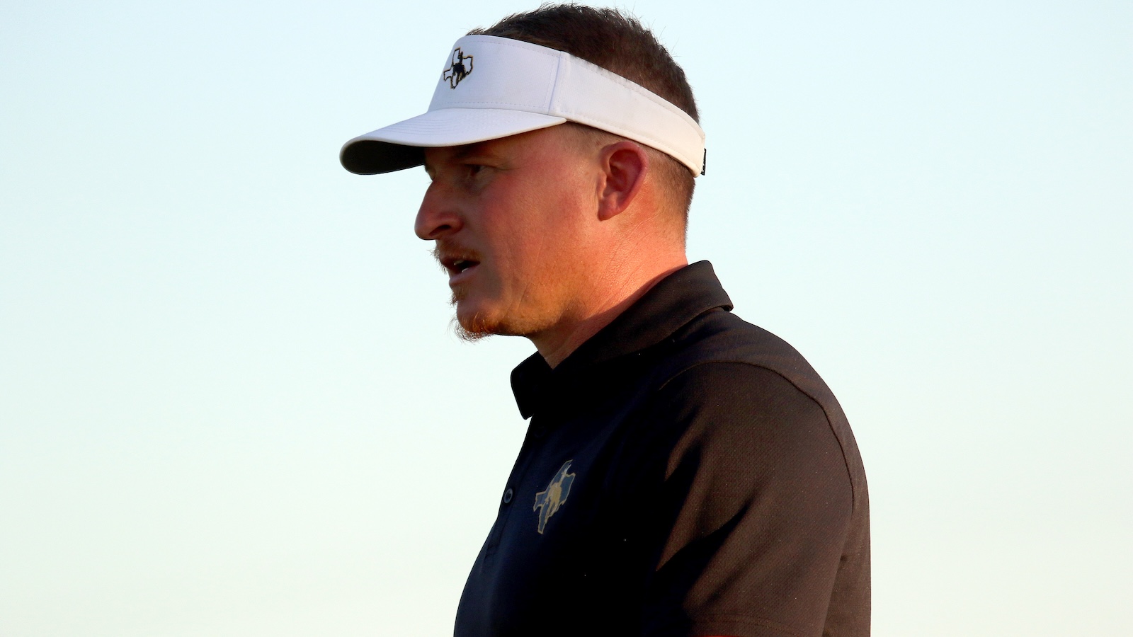 Side profile of a man wearing a white visor and black polo shirt, outdoors at sunset/late afternoon light.