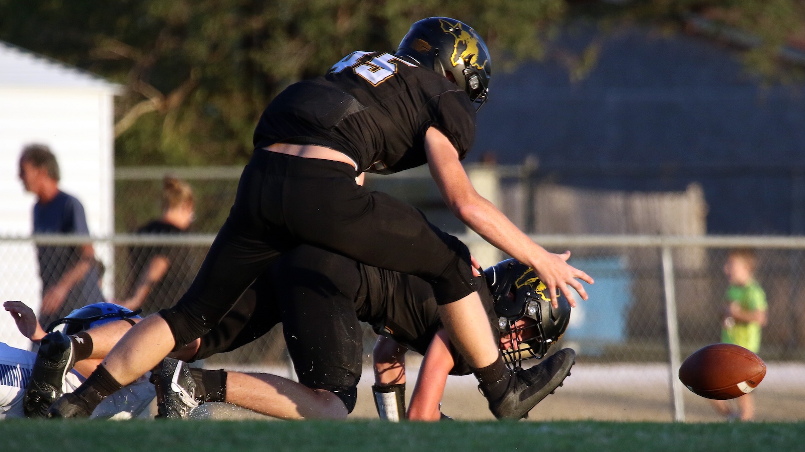 Players in black uniforms tumble on a football field during a tackle as the loose ball rolls nearby on the ground.