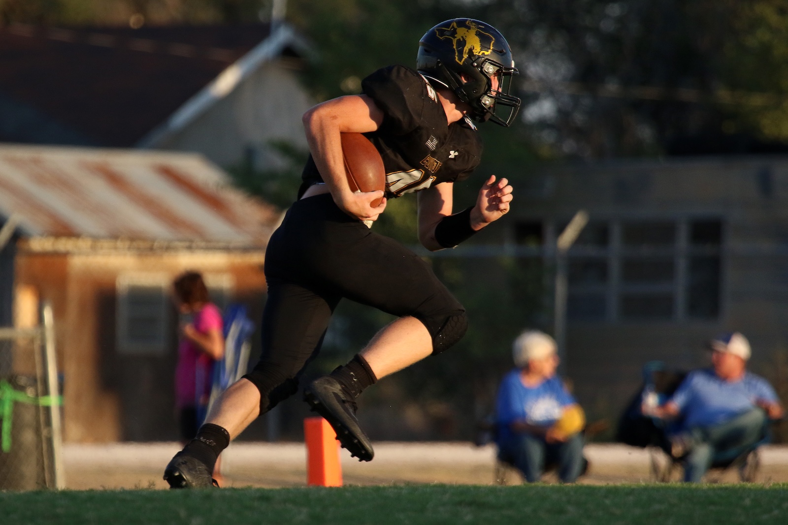 Football player in black uniform chasing the ball while airborne, holding a football against a blurred background of spectators and buildings.