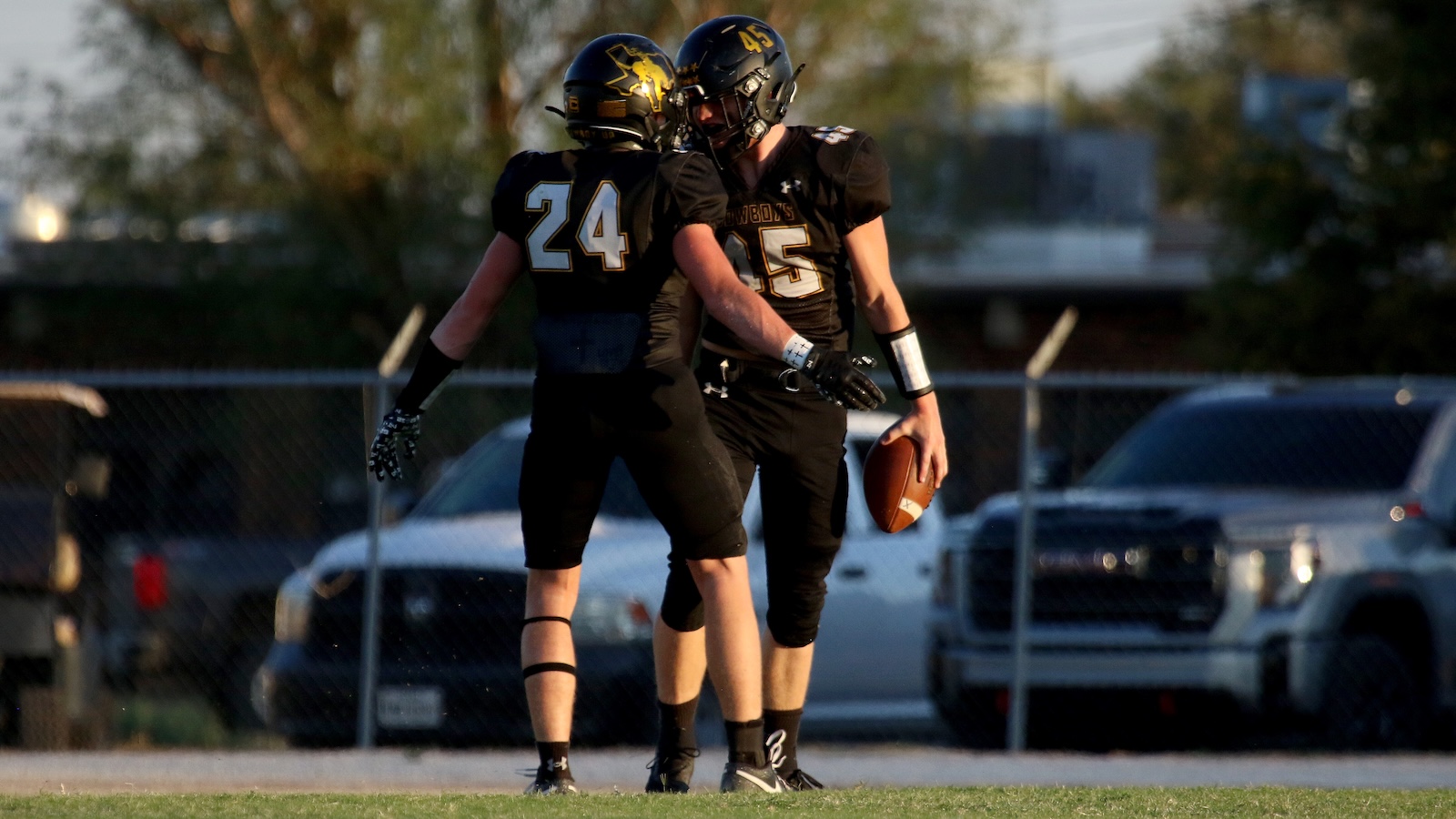 Two football players in black uniforms stand close on the field; one holds a football near his side.
