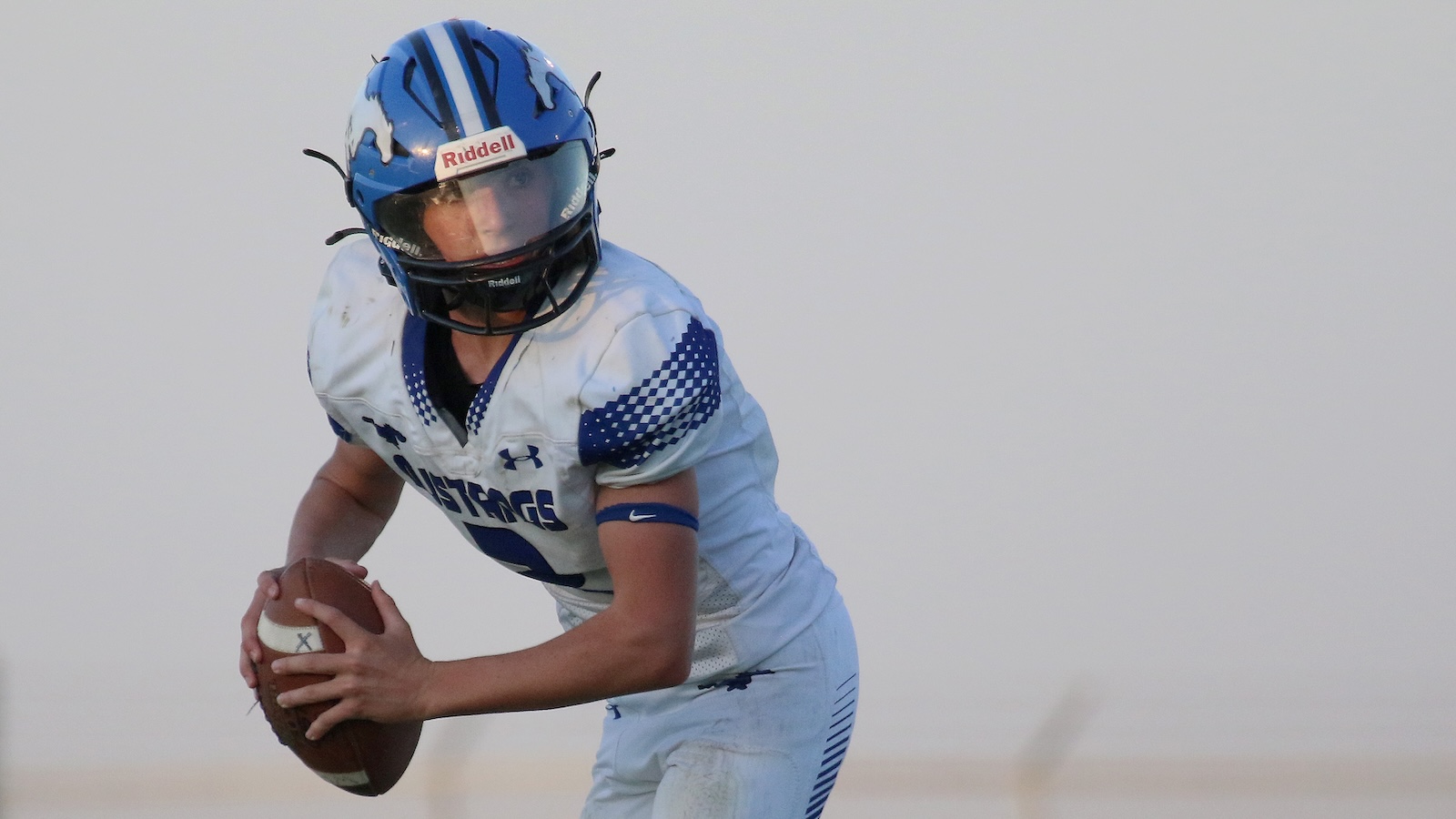 American football quarterback in a white and blue uniform, wearing a blue helmet, holding the ball and scanning the field for an open receiver.