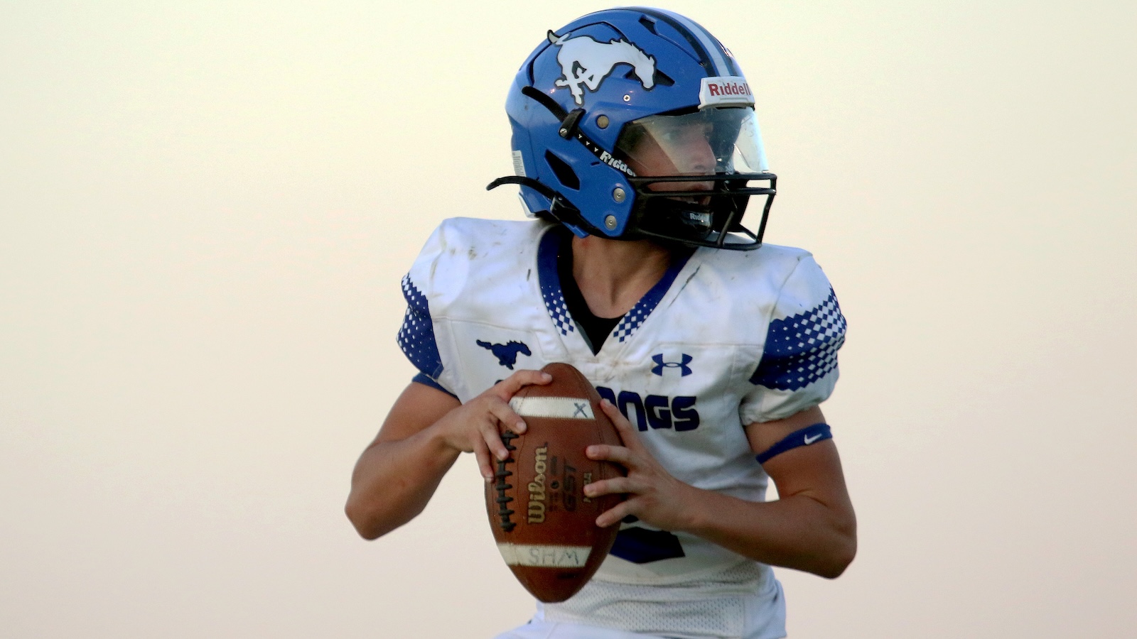 Football player in white and blue uniform holding a football, wearing a blue helmet with a mustang logo.