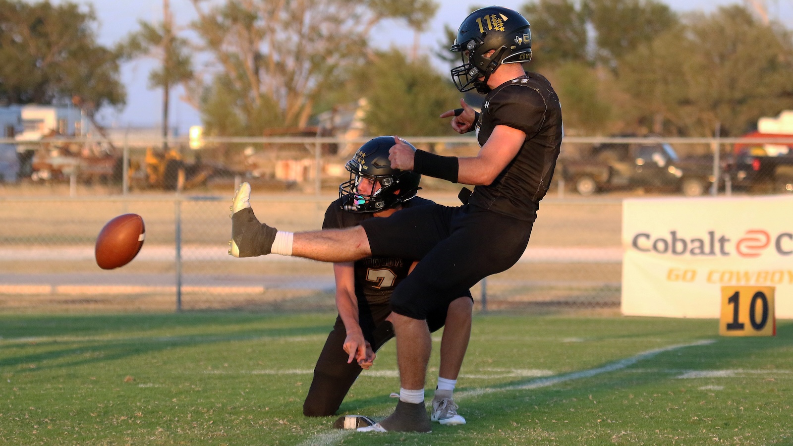 Two football players in black uniforms on a grassy field; the kicker leaps as the ball sails toward the left. Another player kneels behind him, watching the play.