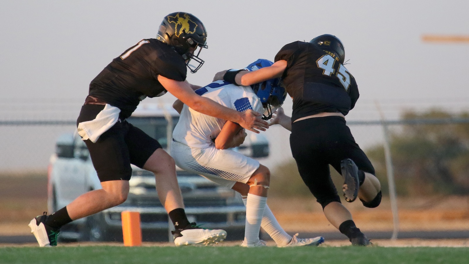 Two football defenders in black jerseys tackle a ball carrier in a white uniform on a grassy field.