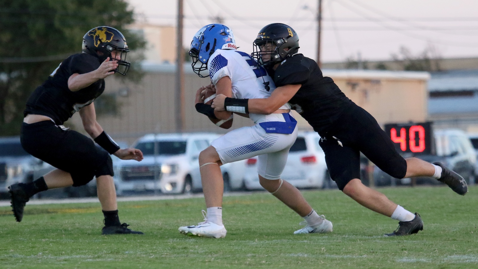 Football game: a player in white uniform holds the ball while facing two opponents in black jerseys tackling him on a grassy field. A digital scoreboard showing 40 is in the background.