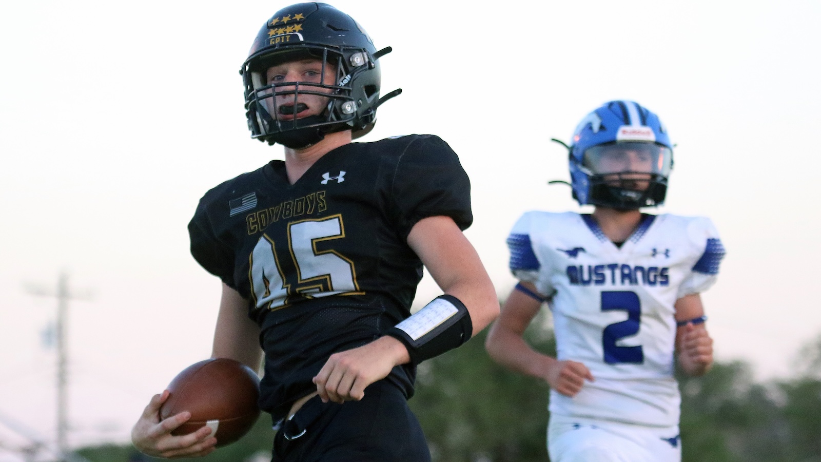 Young football player in a black Cowboys uniform runs with the ball, helmet on, in foreground.